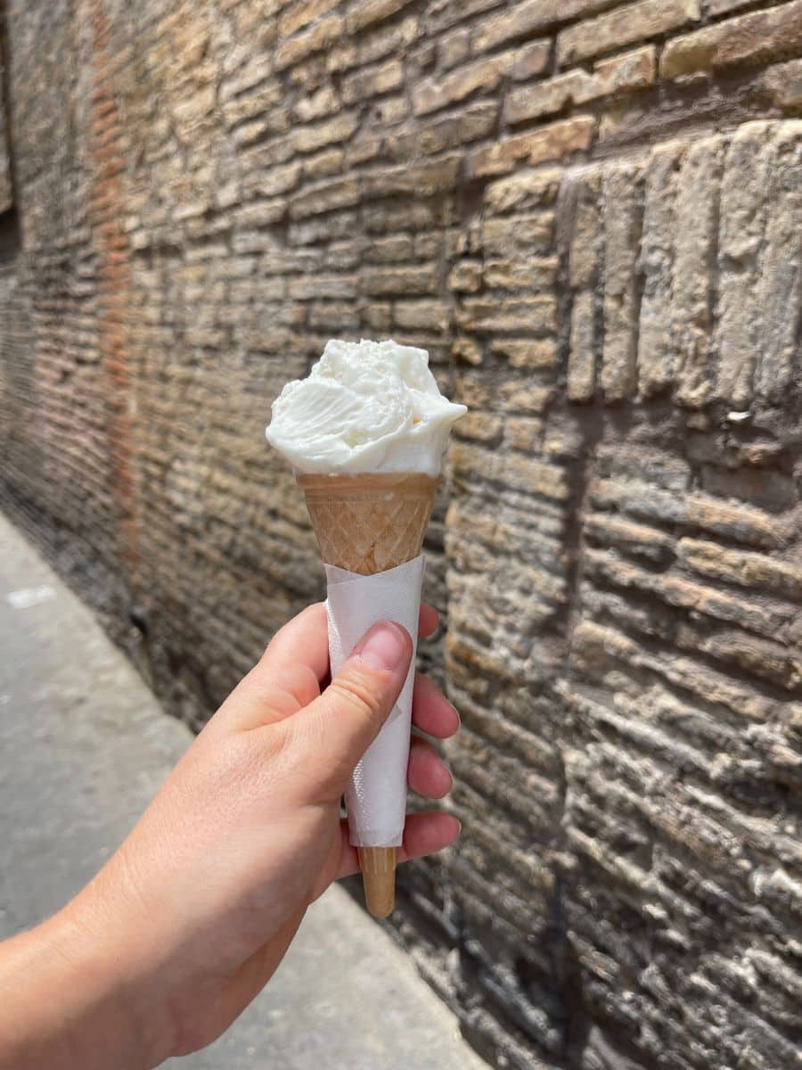 A hand holding a gelato cone with a scoop of creamy white gelato against the backdrop of an ancient brick wall in Rome, Italy. The textured bricks add a rustic charm, contrasting with the smooth, delicious-looking gelato.