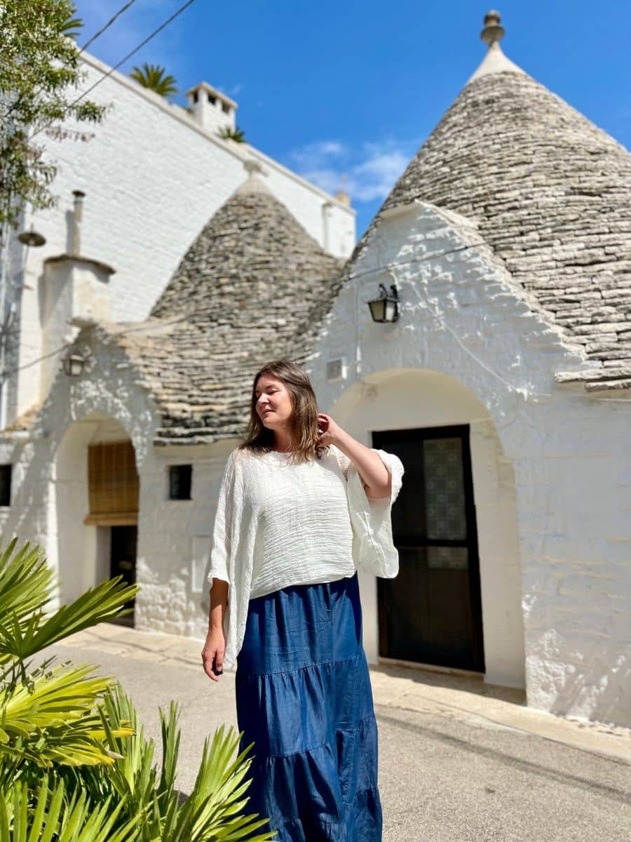 A woman with light skin and long brown hair stands in front of traditional trulli houses in Alberobello, Italy. She is wearing a loose white blouse and a flowing blue skirt, with one hand gently touching her hair. The bright sunlight highlights the whitewashed stone walls and conical roofs of the trulli, contrasting against the vivid blue sky. Green palm leaves frame the lower part of the image, adding a touch of nature to the scene.