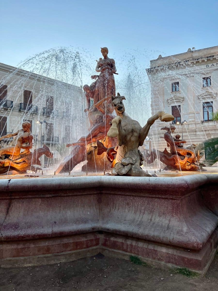 A grand fountain featuring a central statue of a woman standing tall, surrounded by mythological figures, horses, and sea creatures, with water jets spraying in various directions. The sculptures display intricate details, including a striking figure of a rearing horse and a kneeling mythical creature in the foreground. The fountain is set in a European-style square with elegant, historic buildings in the background.