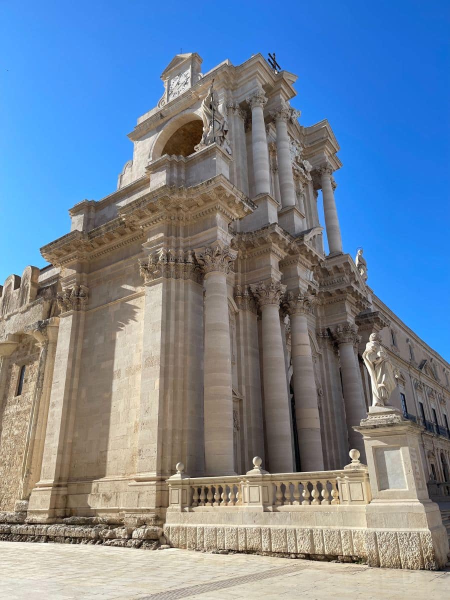 A stunning side view of the Cathedral of Syracuse in Sicily, Italy, showcasing its grand Baroque façade with towering Corinthian columns, intricate stone carvings, and statues. A clock is visible at the top of the structure, adding a historical charm. The bright blue sky provides a striking contrast to the warm, beige stone. A balustrade in the foreground and a statue on a pedestal enhance the architectural grandeur of this UNESCO World Heritage Site.