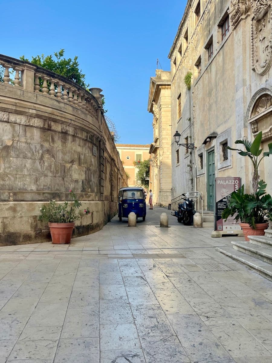 A charming, narrow stone-paved street in Siracusa, Sicily, flanked by historic buildings with aged facades and intricate architectural details. A small, blue three-wheeled vehicle (Ape Piaggio) is parked along the path, adding a quaint touch to the scene. A couple of potted plants and scooters decorate the area, while a pedestrian walks in the background beneath the warm afternoon sunlight. The sky above is clear and vividly blue, emphasizing the serene Mediterranean atmosphere.