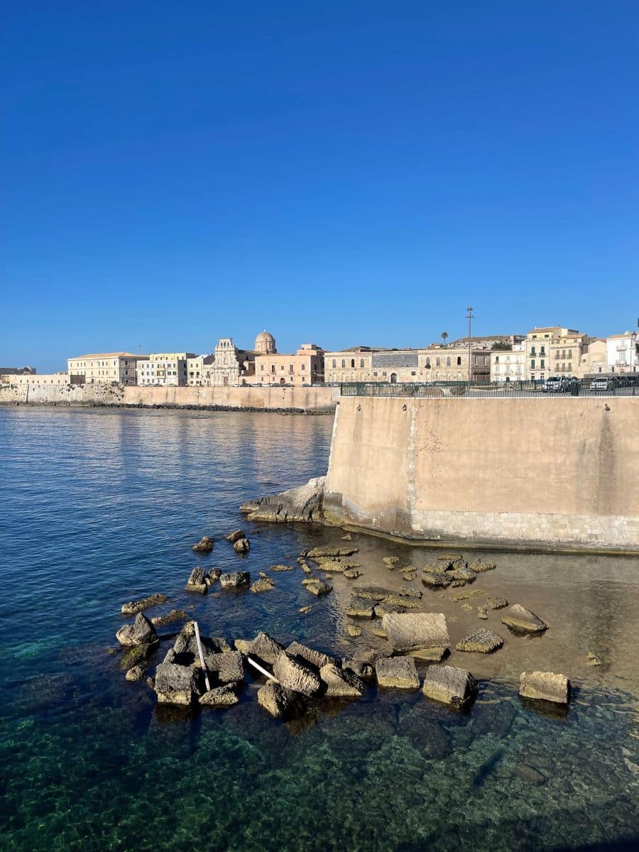 A scenic coastal view of a historic town with beige and pastel-colored buildings along the waterfront. A large stone seawall extends into the clear, blue water, with scattered rocks and remnants of old structures visible in the shallow sea. The sky is bright blue, creating a striking contrast with the calm, transparent water.