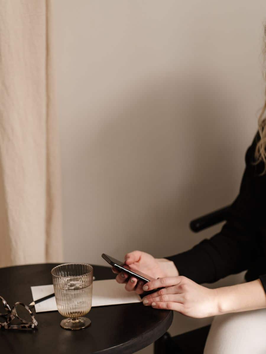 A person sitting at a table, holding a smartphone, with a glass of water, a pen, and a notepad nearby—suggesting communication and connection.