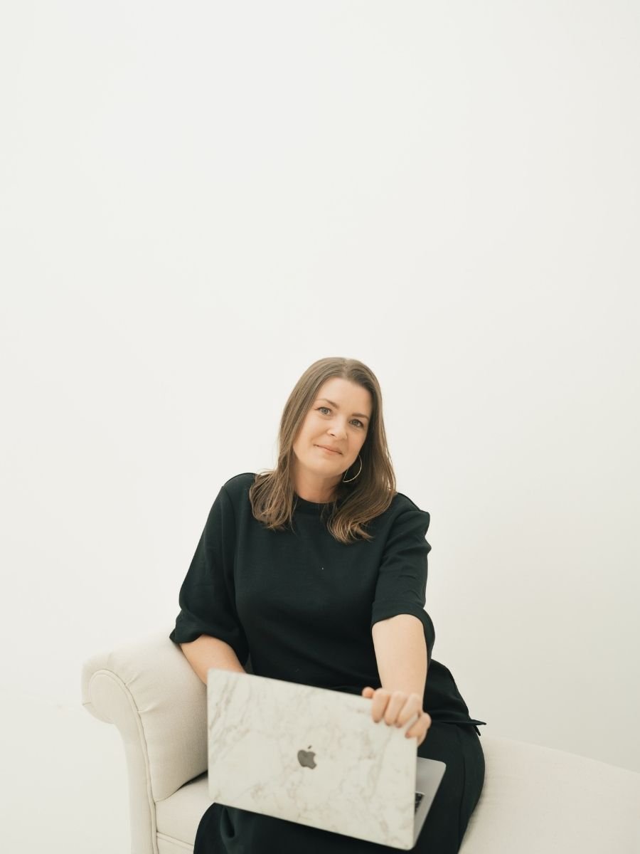 Melissa Byron, founder of A Solo Woman Traveling, seated on a light couch holding a laptop while working as a travel advisor and entrepreneur.