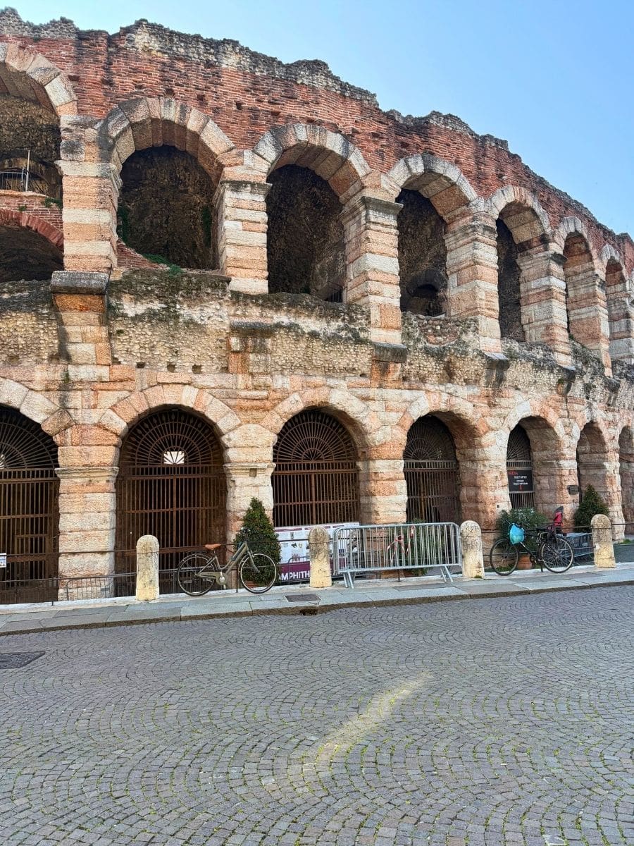 Ancient Roman amphitheater, the Verona Arena, with weathered pink and white stone arches, barred gates at ground level, and bicycles parked along the cobblestone street in front.