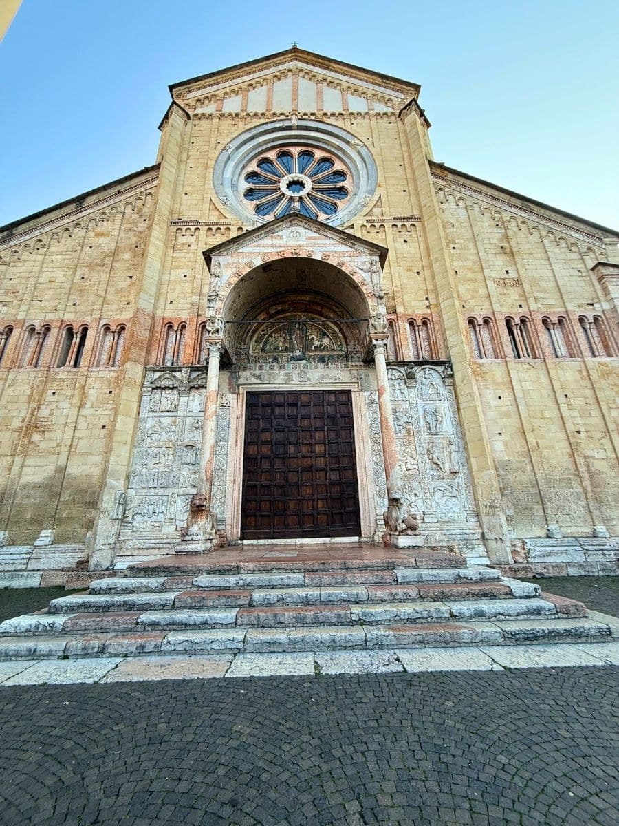 A close-up view of the ornate entrance to the Basilica of San Zeno in Verona, Italy, showcasing the intricately carved stone doorway, a large wooden door, and the detailed arch above adorned with frescoes and sculptural elements. The iconic rose window sits prominently above the entrance, framed by the Romanesque fa&ccedil;ade of warm yellow and red stone.