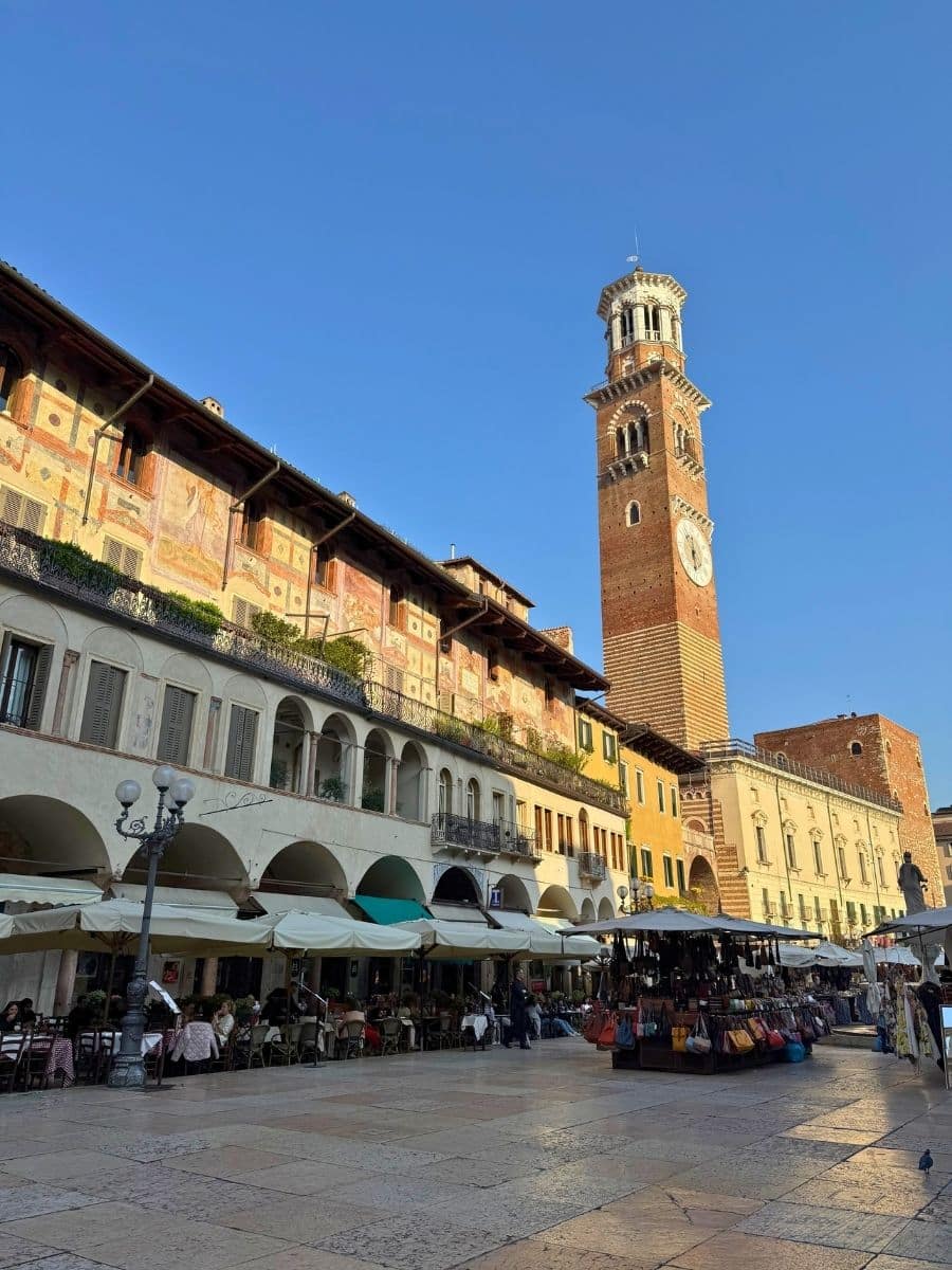 A bustling piazza in Verona under a clear blue sky, showcasing historic buildings with faded frescoes and arched walkways lining the left side. The prominent Torre dei Lamberti, a tall brick tower with a clock face and white accents, rises on the right, casting a shadow across the paved square where outdoor cafes with white umbrellas and market stalls are active with people.