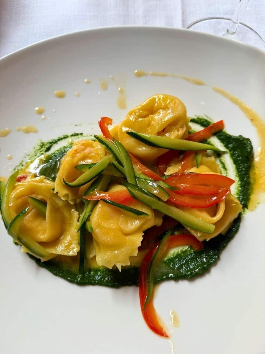 A beautifully plated dish of tortellini pasta, served with thinly sliced red bell peppers and zucchini on a vibrant green sauce, possibly made from herbs or spinach. The tortellini is arranged artfully on a white plate, and the dish is lightly drizzled with a golden sauce, creating a colorful and appetizing presentation perfect for a fine dining experience in Italy.