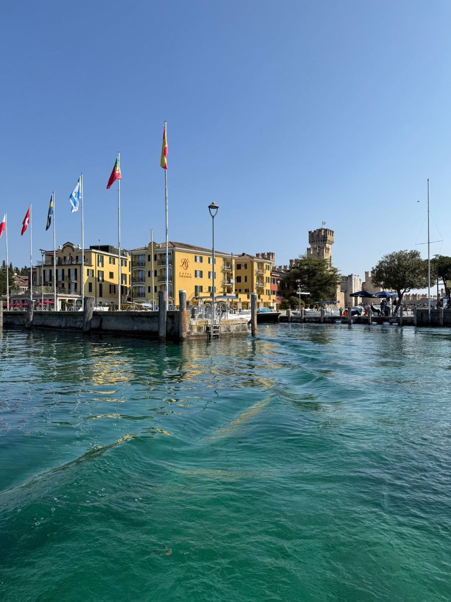 A picturesque waterfront view in Sirmione, Italy, showing the shimmering turquoise waters of Lake Garda under a clear blue sky. Multiple national flags are raised on tall poles along the pier. Behind the flags, the yellow fa&ccedil;ade of Hotel Sirmione e Promessi Sposi is visible, flanked by charming historic buildings and the recognizable medieval tower of Scaliger Castle in the background. The scene captures a sunny, peaceful atmosphere typical of this scenic lakeside town.