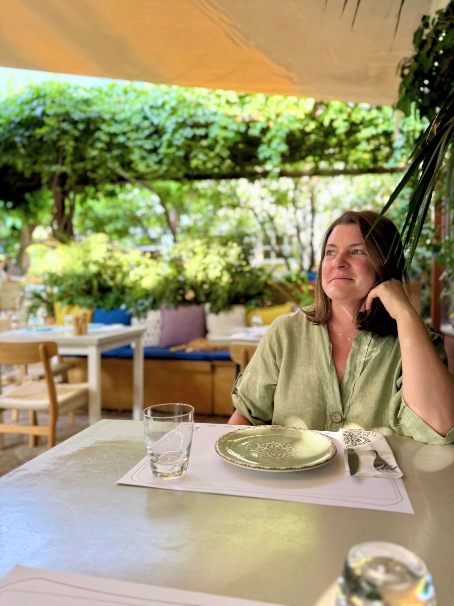 Melissa in a green blouse sits at an outdoor restaurant in Hydra, Greece, surrounded by lush greenery and colorful cushions, gazing thoughtfully with a soft smile.