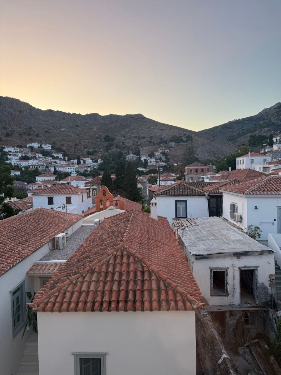 Sunset over the hillside homes of Hydra, Greece, featuring classic whitewashed buildings with terracotta rooftops nestled against the mountainous terrain. The sky fades from golden to lavender as evening approaches.