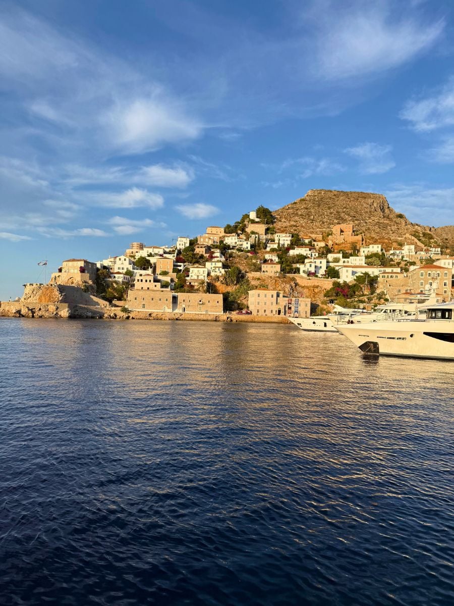 Scenic coastal view of Hydra, Greece at golden hour, featuring traditional stone houses built on a hillside overlooking the deep blue Aegean Sea with a few docked yachts in the foreground.