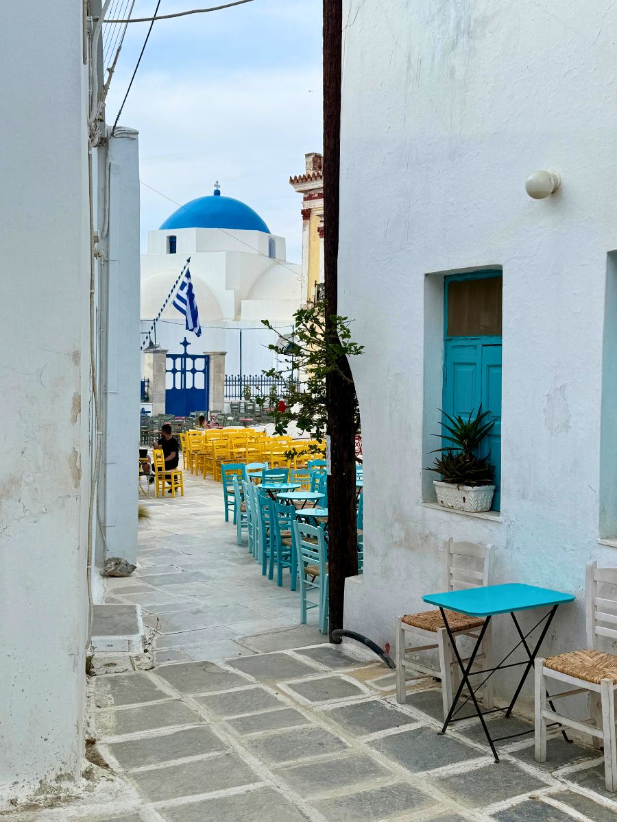 Narrow alley in Serifos, Greece, opening to a lively courtyard filled with vibrant yellow and turquoise café chairs, with a traditional Greek church and its iconic blue dome in the background.