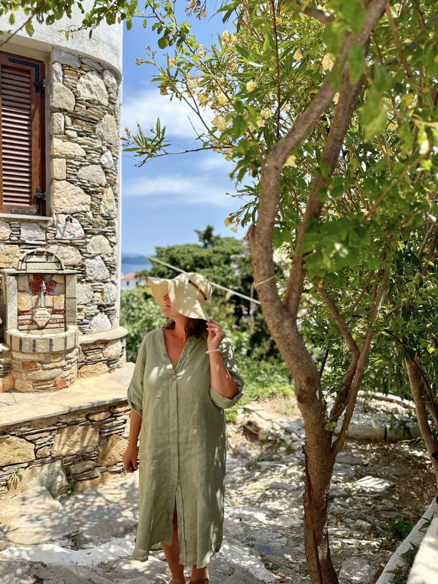 Melissa in a loose green dress and sunhat standing under leafy trees next to a rustic stone building in Alonissos, Greece.