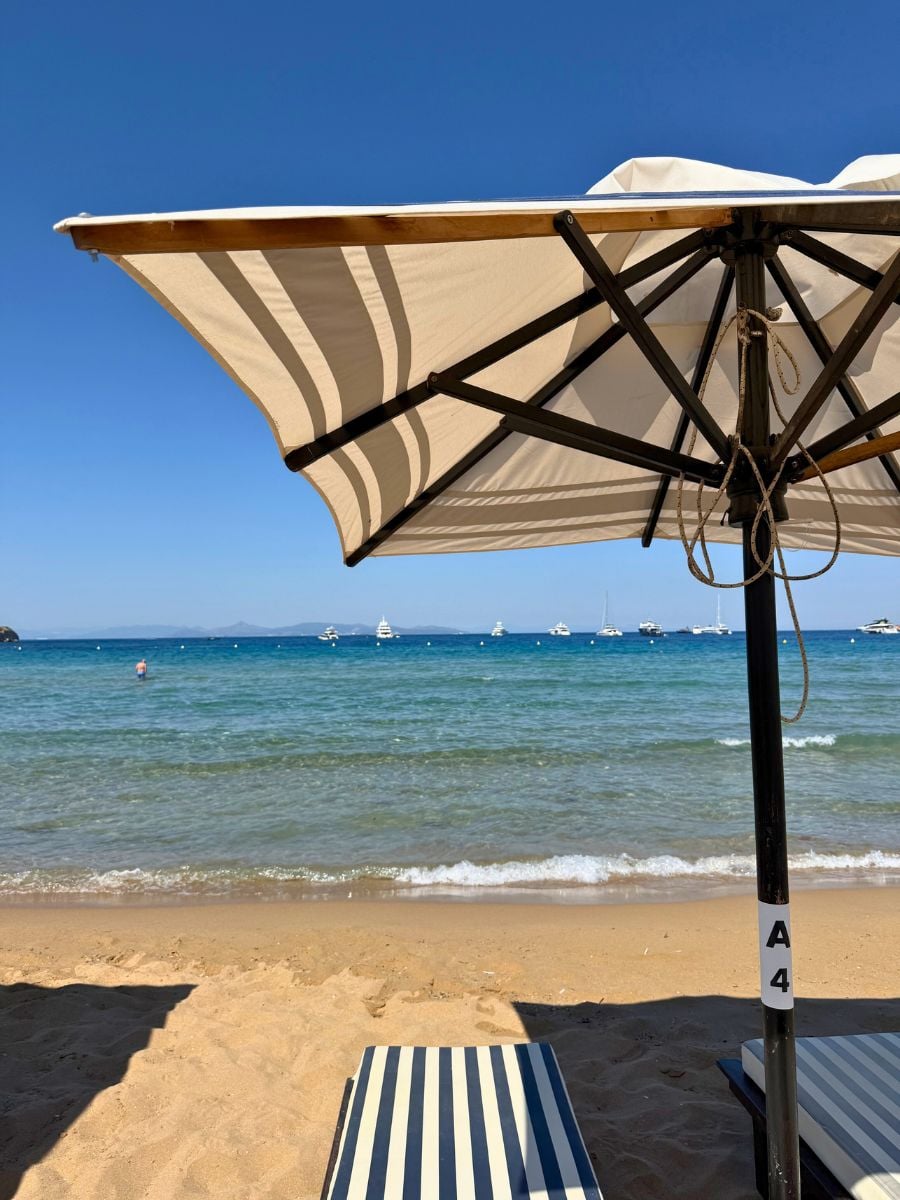 View from a shaded beach chair under a striped umbrella, looking out at the clear blue waters and anchored boats off the coast of Naxos, Greece.