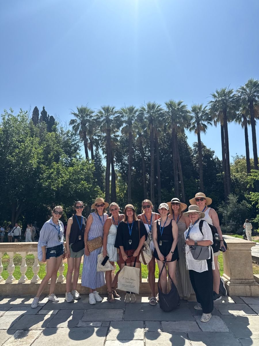 A group of women pose together under the bright sun in Athens' National Garden, surrounded by lush greenery and tall palm trees.