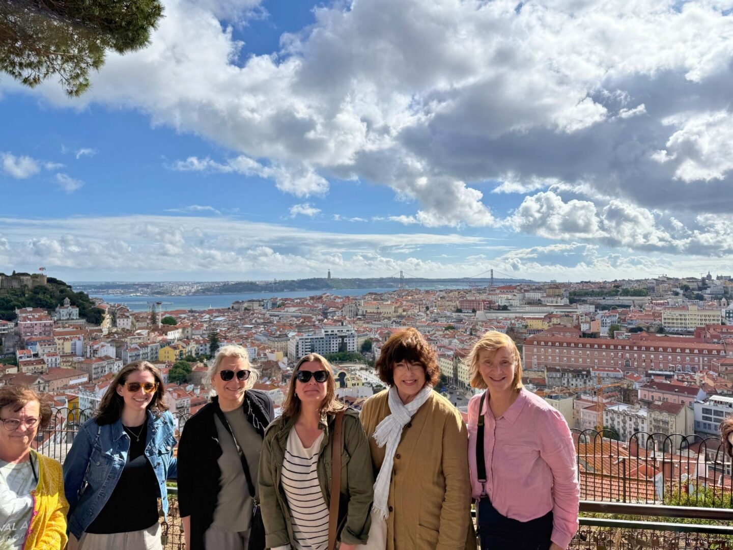 A group of six women smiling in front of a scenic overlook of Lisbon, Portugal, with red-tiled rooftops, the Tagus River, and the 25 de Abril Bridge in the background. The photo captures a bright, partly cloudy day during a small group women’s trip.