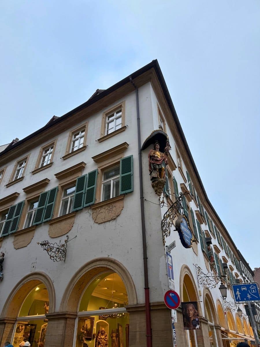 Corner view of a historic building in Bamberg with green window shutters, arched shop windows, ornate wrought-iron signs, and a statue of a saint on the upper façade under an overcast sky.