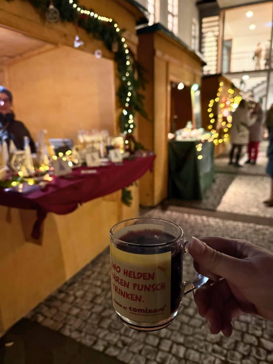A hand holding a mug of hot punch at a festive Christmas market stall in Bamberg, with twinkling fairy lights and decorated wooden booths in the background.
