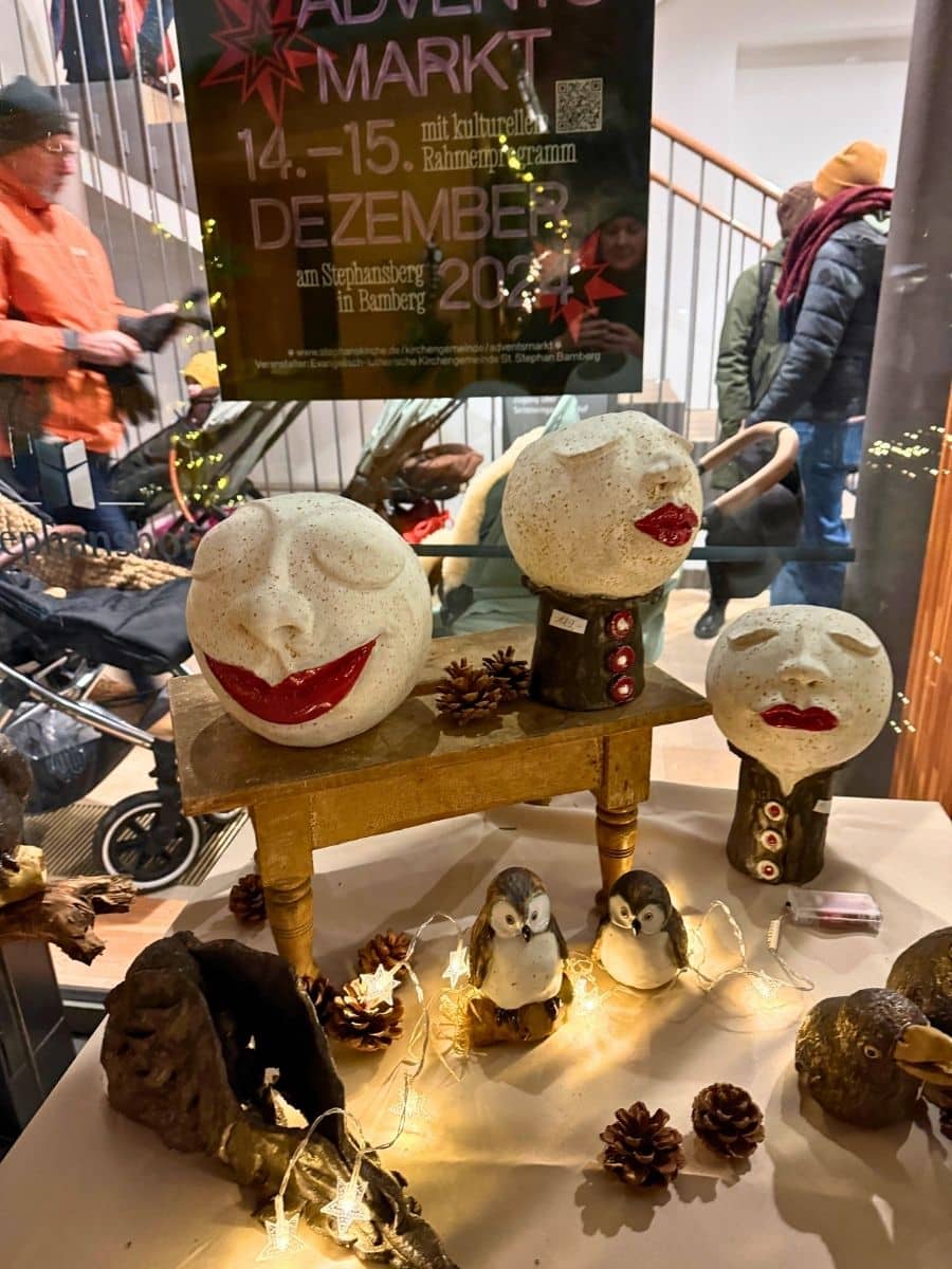 Quirky handcrafted ceramic faces and animal figurines displayed with pinecones and fairy lights at Bamberg’s Advent Market, with a poster showing the event dates in the background.