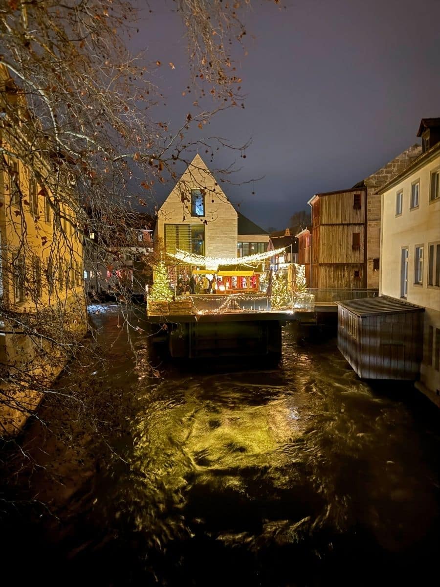 Beautifully lit wooden pavilion floating on the river in Bamberg at night, adorned with Christmas lights and surrounded by historic buildings.