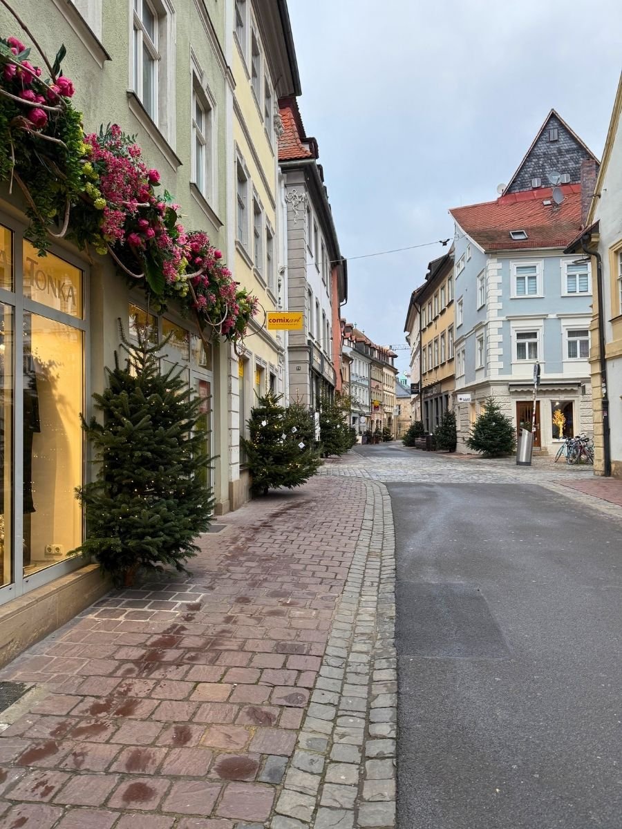Festively decorated street in Bamberg with Christmas trees and garlands, showing pastel facades and boutique shops including one named "Tonka.