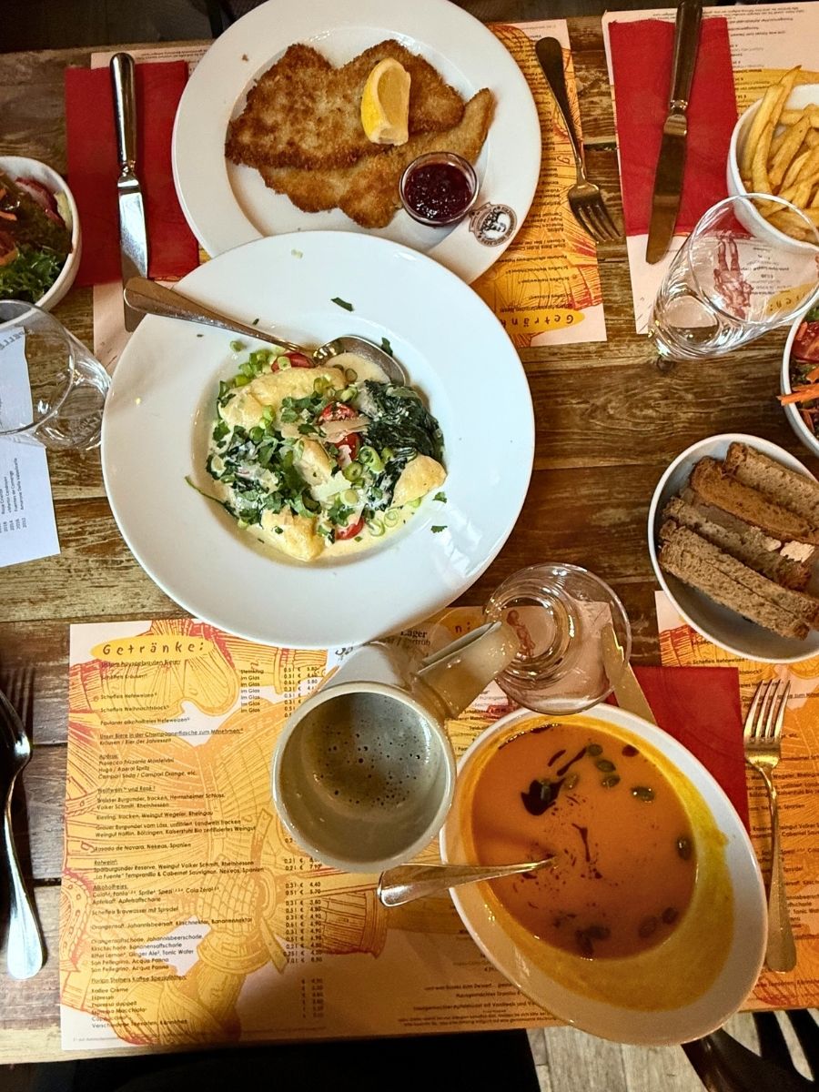 Overhead view of a rustic wooden table set with traditional German dishes, including schnitzel with lemon, creamy vegetable dish, pumpkin soup, dark bread slices, salad, and beer.