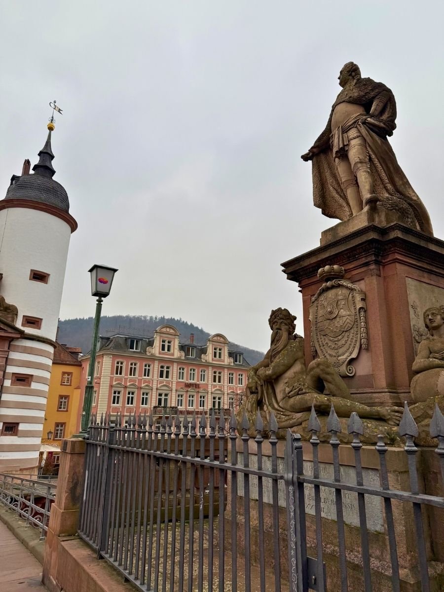 Statue of a historical figure in a cape atop a monument on the Old Bridge, with a pink Baroque-style building and hilly landscape in the background.