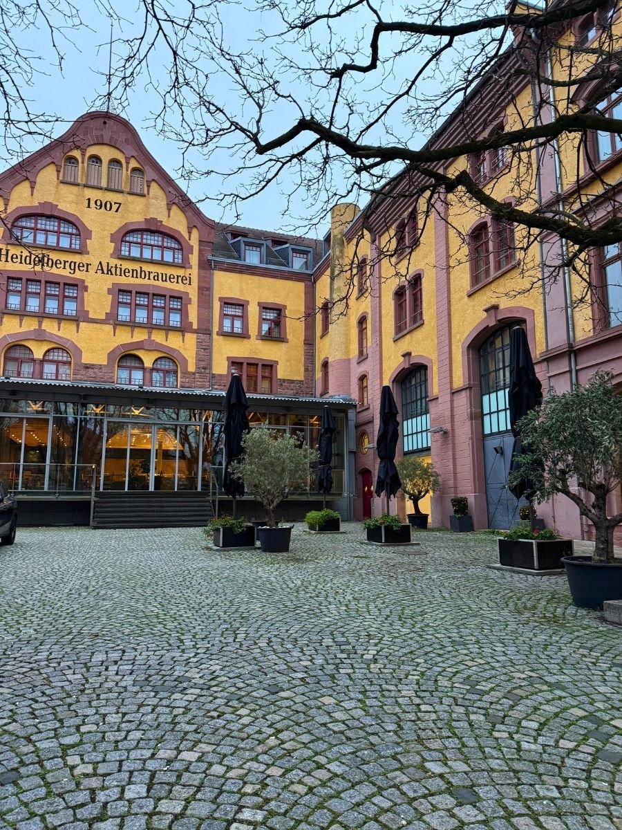 Historic yellow building of Heidelberger Aktienbrauerei with cobblestone courtyard and potted trees.