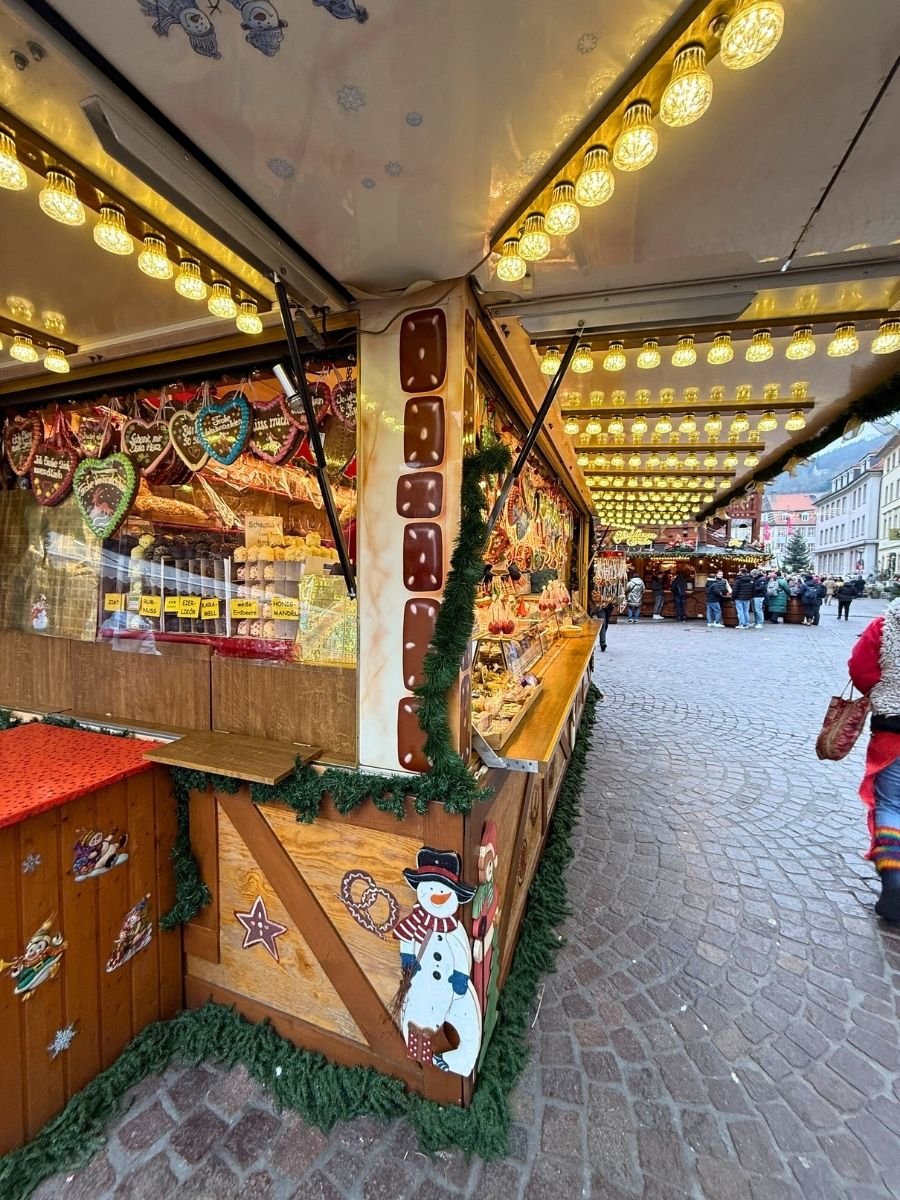 Gingerbread and candy stall at a Christmas market, decorated like a gingerbread house with festive lights and a cheerful snowman painting.