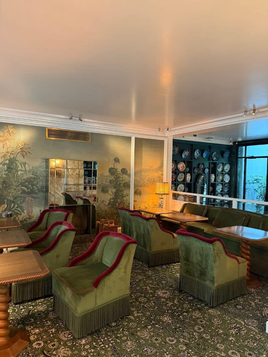 Vintage-style seating area at Hotel Eldorado Paris with plush green armchairs, wooden tables, and painted walls with scenic Asian-inspired murals.