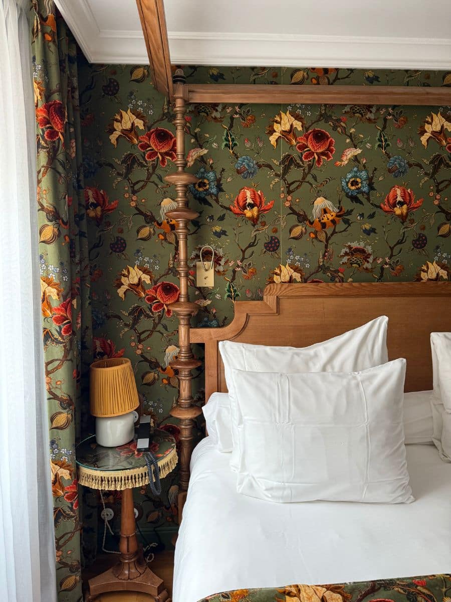 Cozy bedroom at Hotel Eldorado Paris with white pillows, wooden bed frame, and floral wallpaper in rich green and red tones.