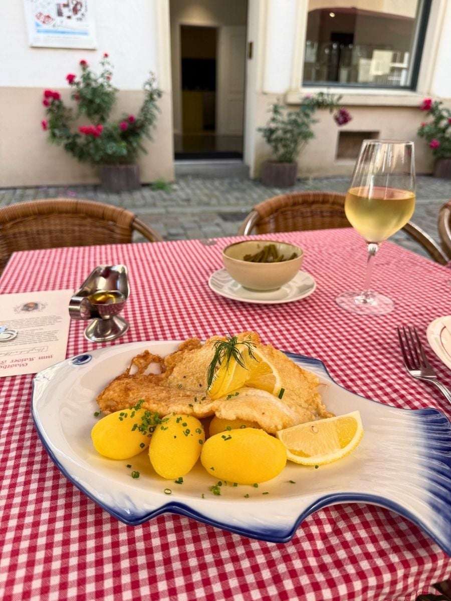 A classic North German dish served outdoors in Bremen&rsquo;s Schnoor Quarter &mdash; golden fried fish with boiled potatoes, lemon, and a glass of white wine. A cozy street-side dining experience on a red-checkered tablecloth.