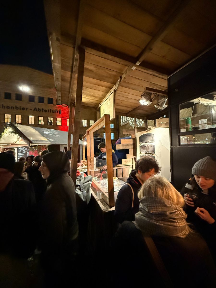 Crowds gathered at a busy wooden food stall at a Berlin Christmas market at night. People are bundled in winter coats and scarves while vendors prepare food behind the counter.