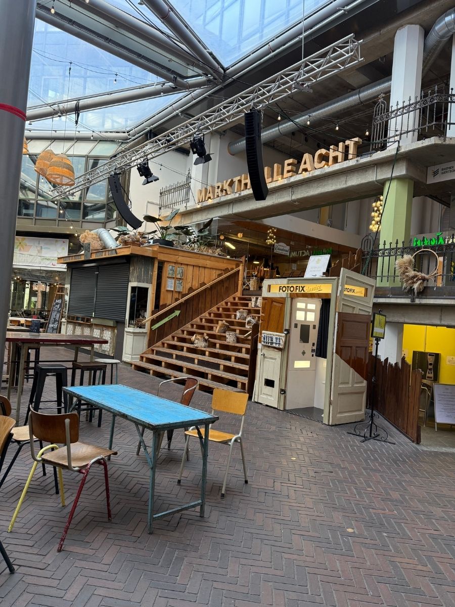 Interior of Markthalle Acht in Bremen, Germany, featuring rustic wooden stalls, tables and chairs for visitors, and a staircase leading to the upper level with the large illuminated sign "MARKTHALLE ACHT.