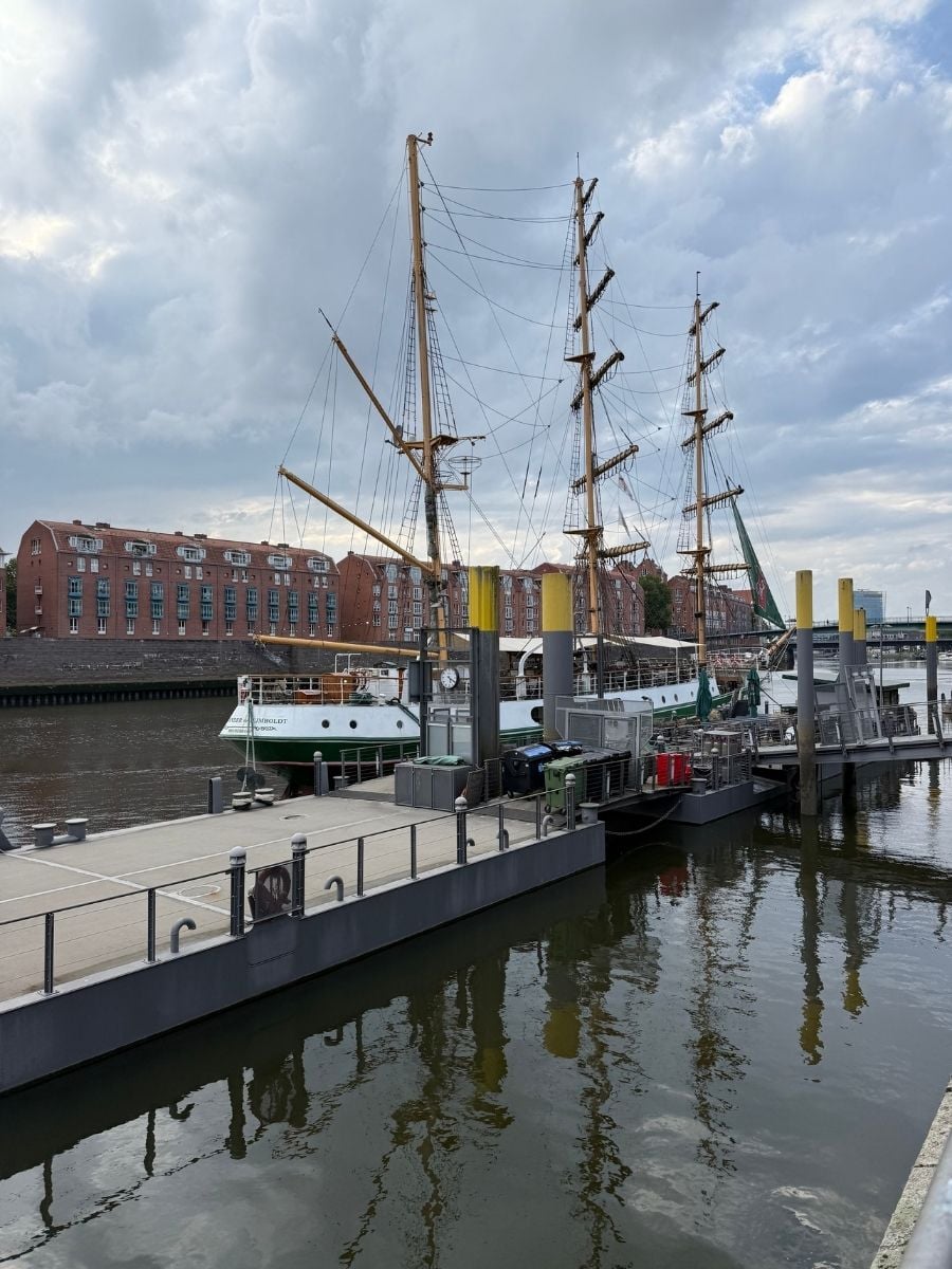 Majestic sailing ships docked along the Weser River in Bremen, with their tall masts rising high against a dramatic cloudy sky.