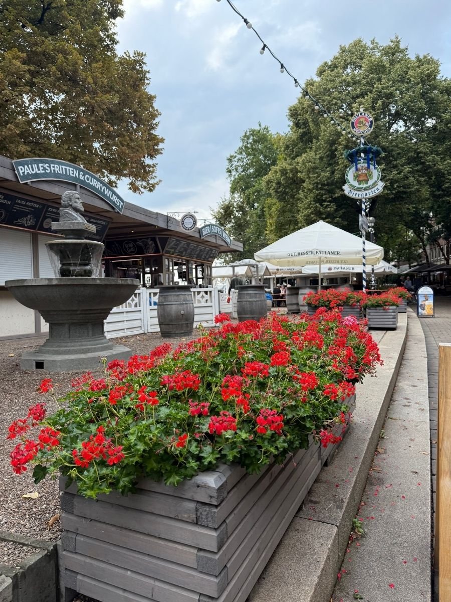 Rows of vibrant red flowers line a walkway at a lively Biergarten in Bremen. Behind them, signs for currywurst and beer stalls can be seen, creating a colorful and inviting scene for food lovers.