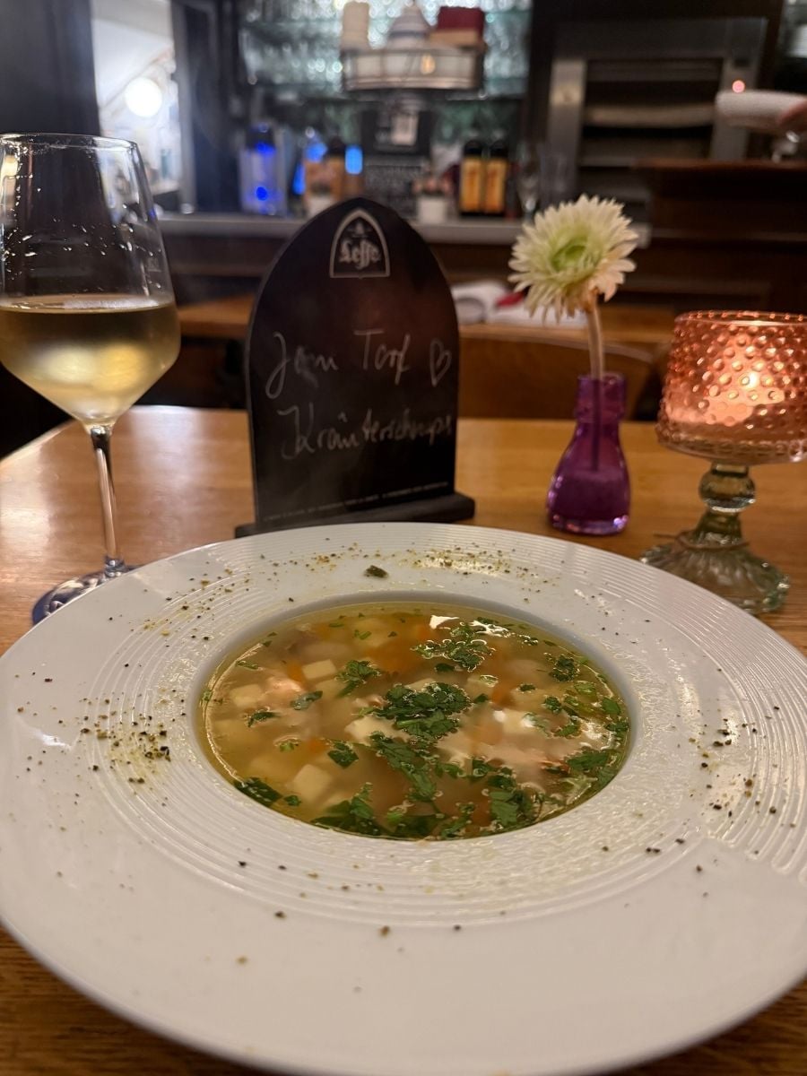 A delicious bowl of soup garnished with herbs is served alongside a glass of white wine in a Bremen restaurant. The setting is warmly lit with a candle and small flower vase, creating the perfect cozy dining experience.