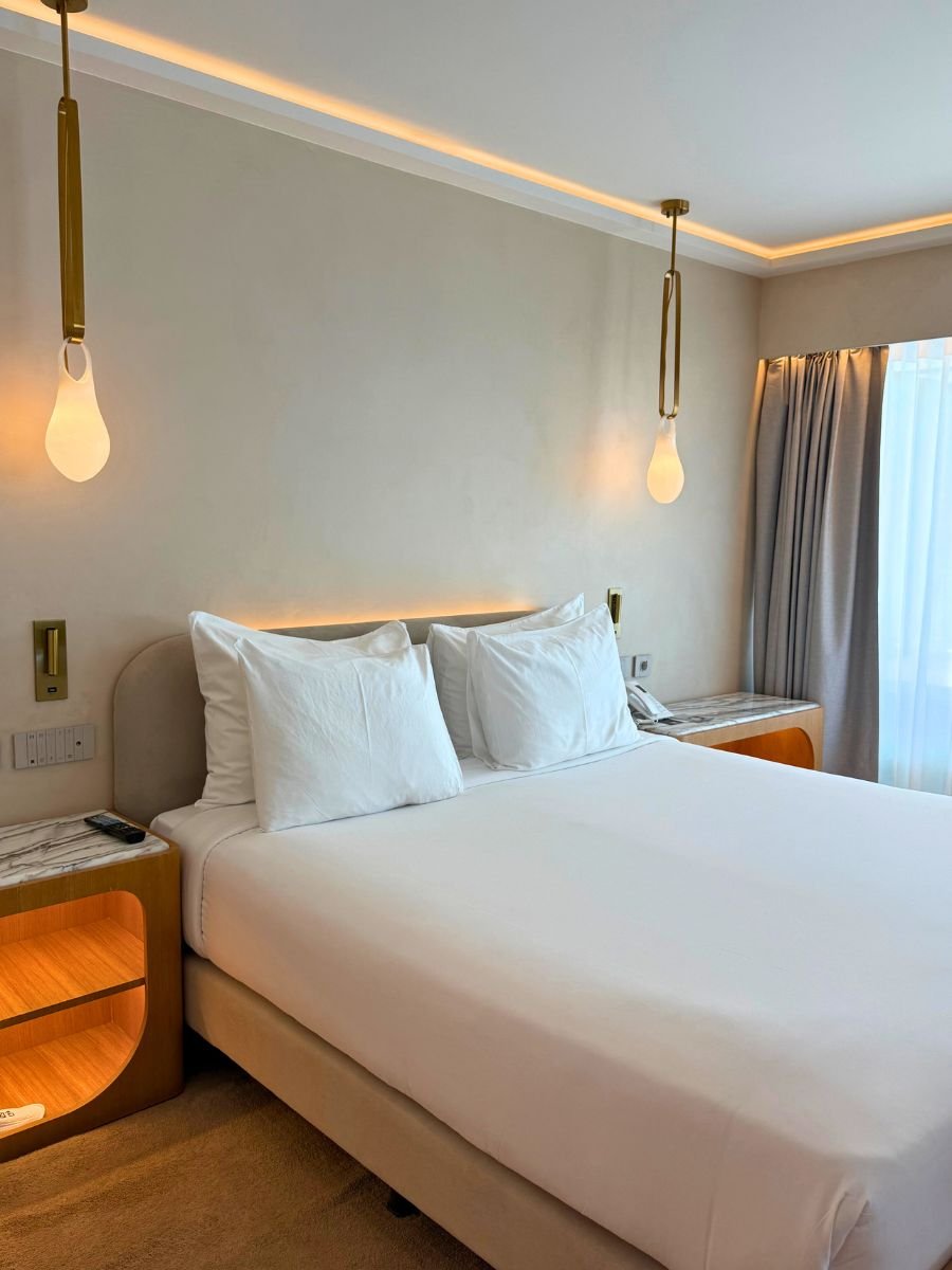 A neatly made double bed with crisp white linens, flanked by marble-topped wooden nightstands and pendant lights, in a softly lit hotel room.