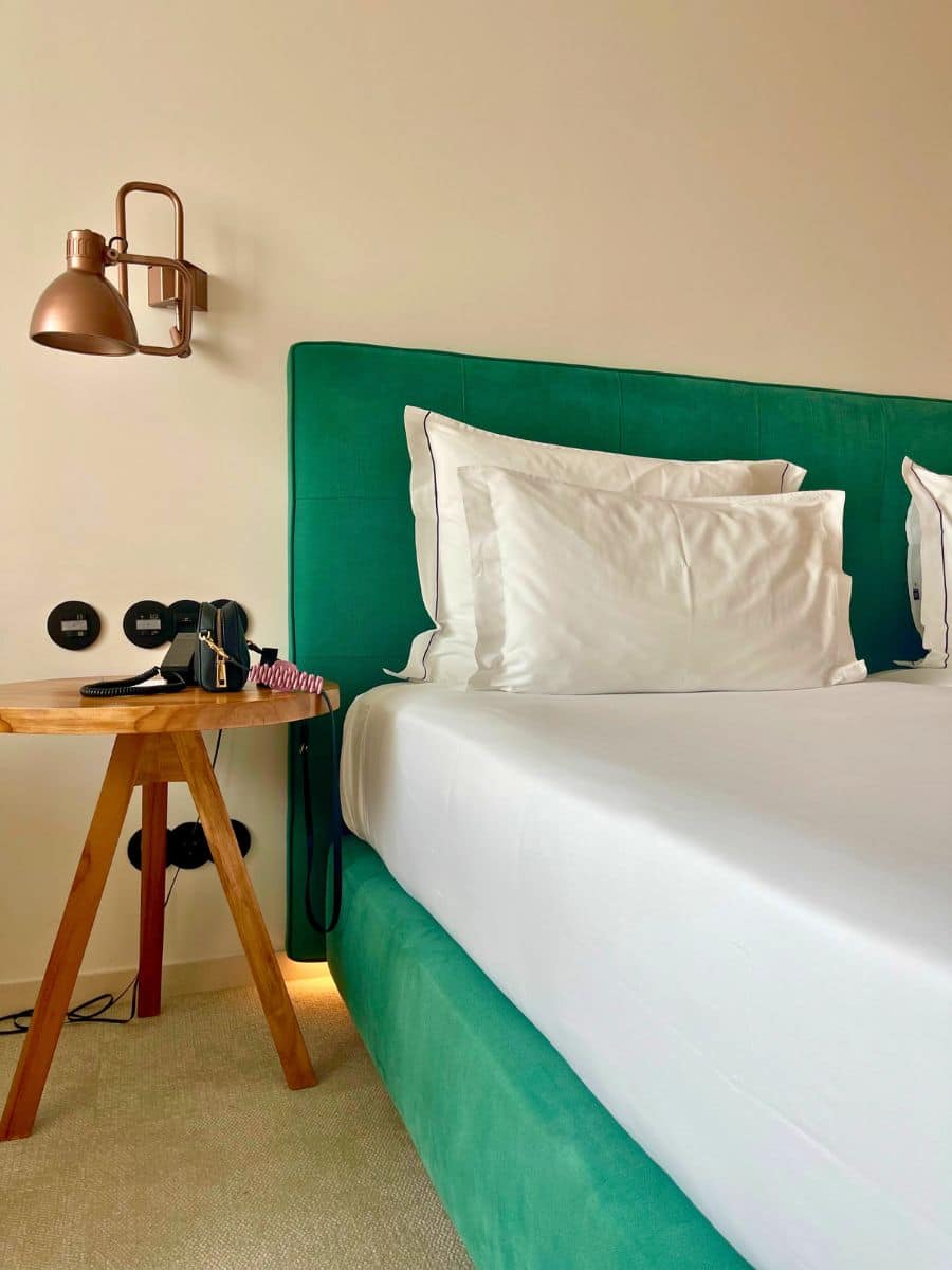 Green velvet headboard, crisp white linens, and wooden side table with copper wall lamp at 3HB Faro.