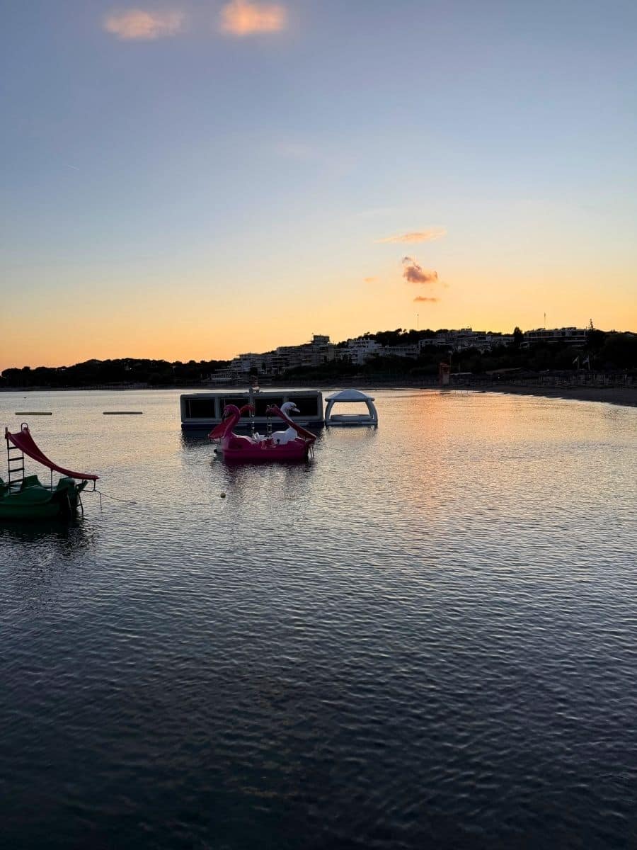 A tranquil sunset over the sea in Milos, with pedal boats and floating platforms silhouetted against the pastel sky.