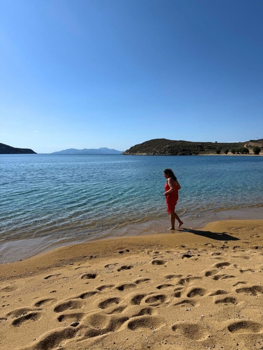 Woman in a red dress walking barefoot along the shoreline of a sandy beach in Milos, with turquoise water and hills in the distance.