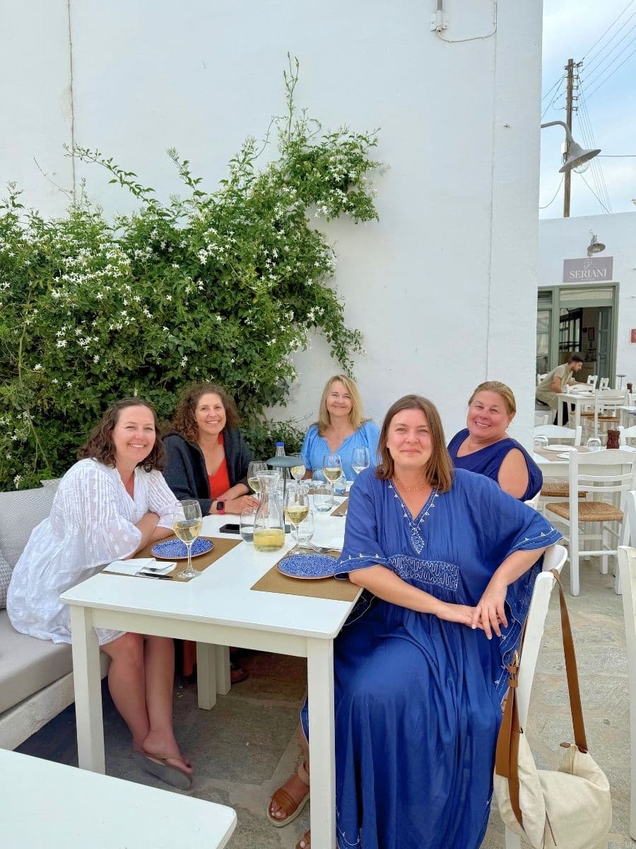 Group of women seated at an outdoor restaurant table in Milos, enjoying drinks and smiling together against a whitewashed wall with greenery.
