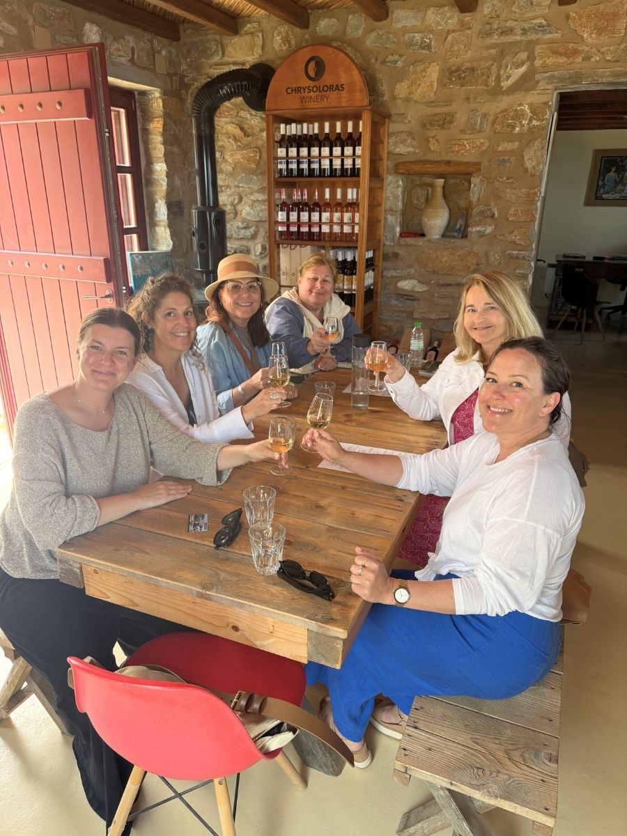Group of six women enjoying wine tasting inside Chrysoloras Winery in Milos, seated at a rustic wooden table.