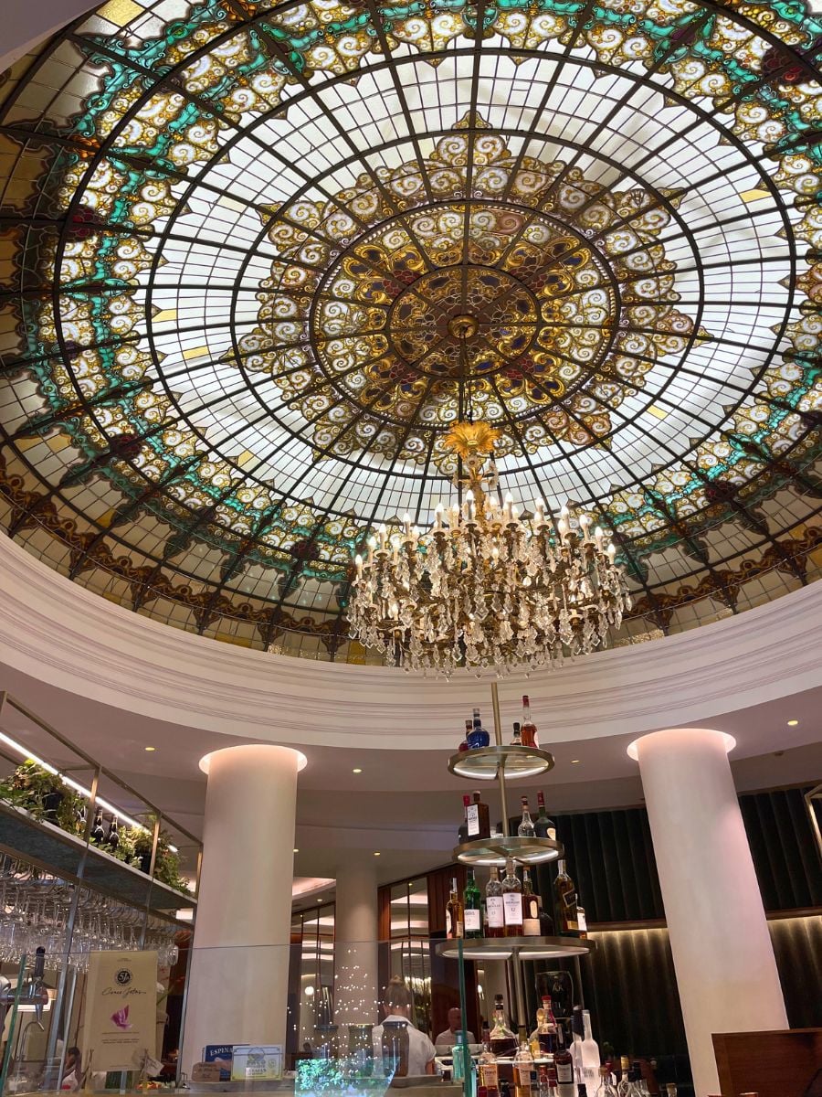 Elegant bar area with a crystal chandelier and a large stained glass dome ceiling in a Seville hotel.