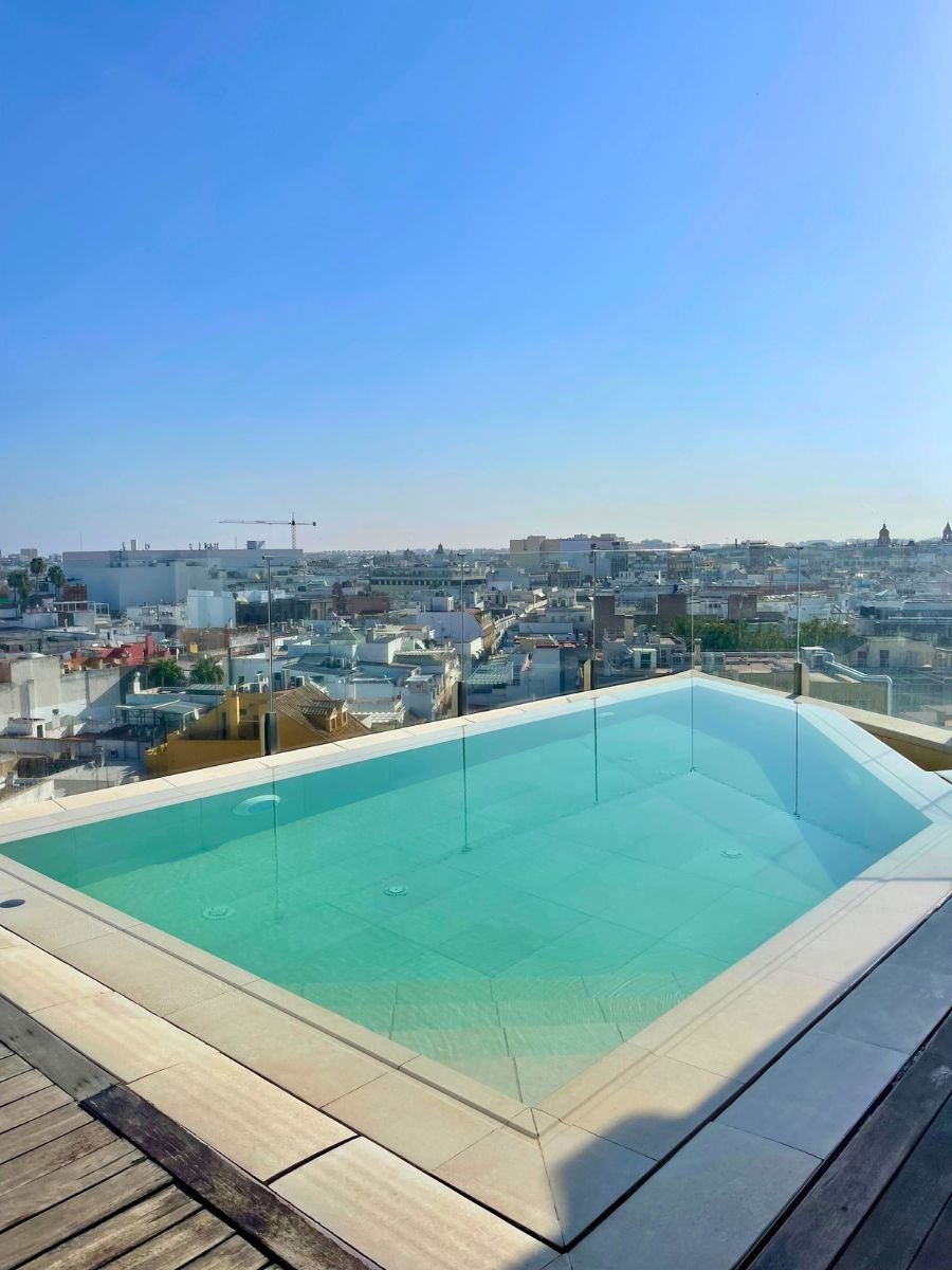 Small rooftop pool with glass panels overlooking Seville&rsquo;s skyline under a clear blue sky.