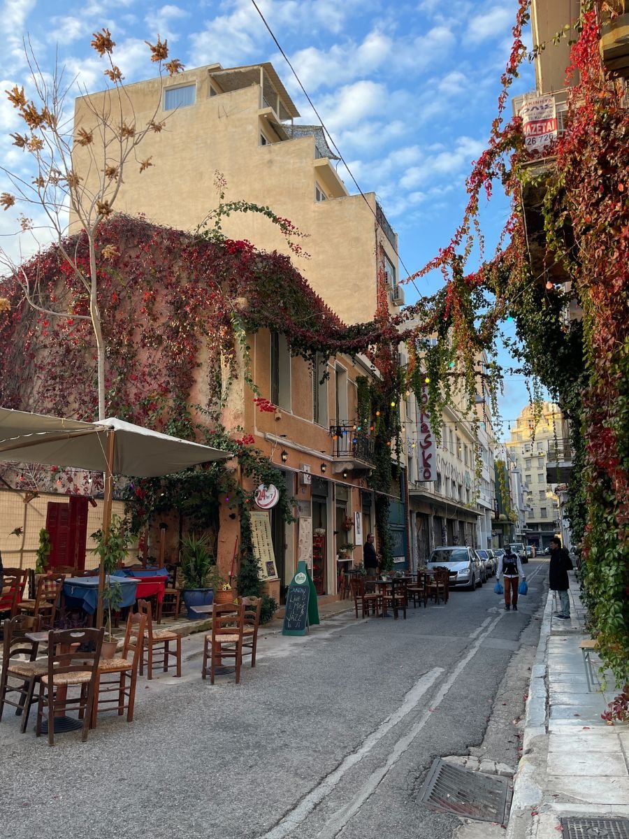 Charming narrow street in Athens lined with outdoor seating, overgrown vines, and cozy local restaurants, under a partly cloudy sky.