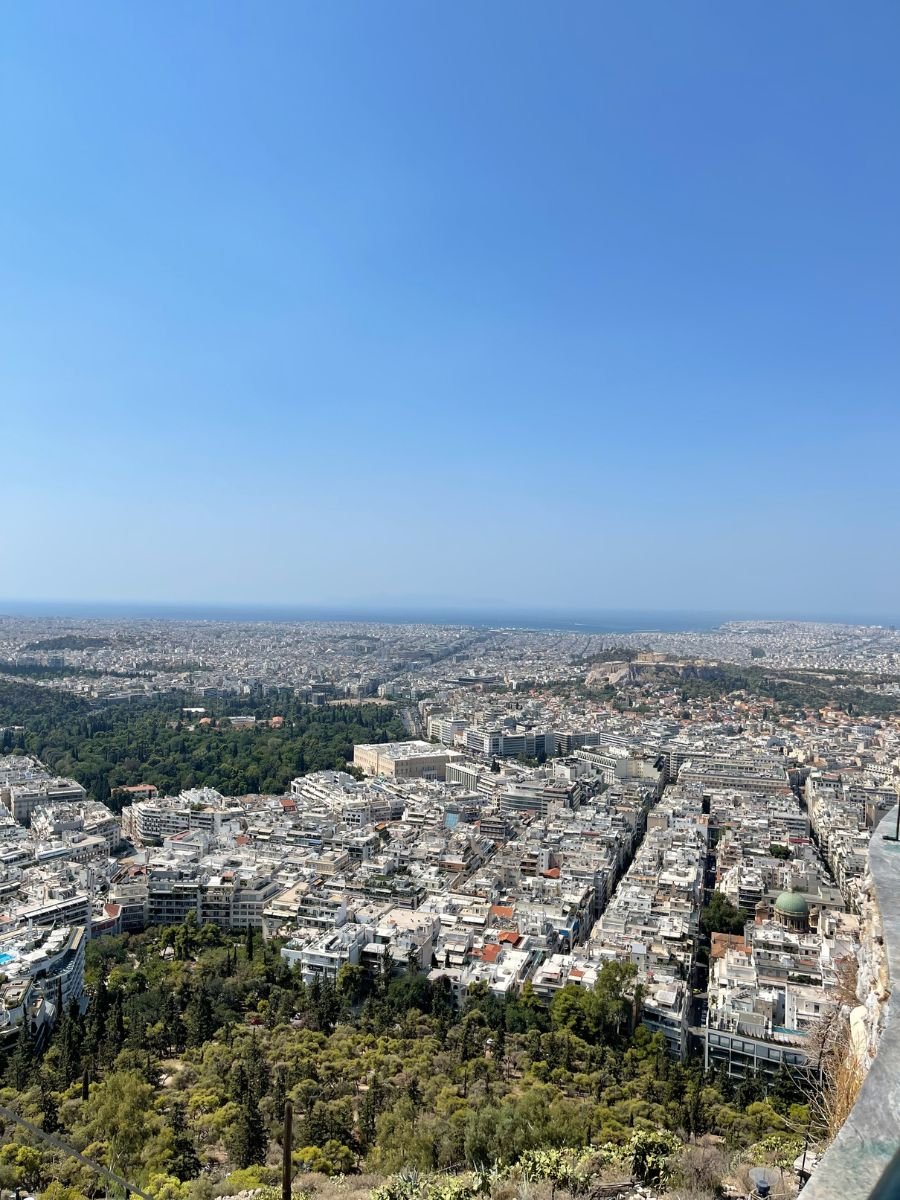 Sweeping aerial view of Athens stretching to the Aegean Sea, with city blocks and green parks under a clear sky.