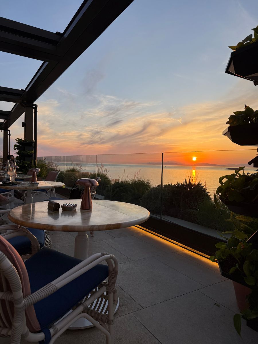 Romantic sunset view from rooftop restaurant in Athens, with marble tables, cozy seating, and golden sun setting over the sea.
