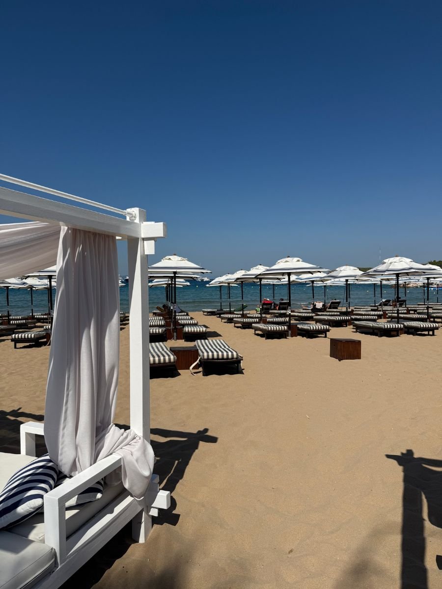 Elegant white cabana and striped loungers on the sandy shore of Astir Beach in Athens, Greece, facing the clear blue Aegean Sea under a sunny sky.